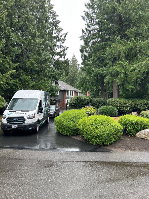 A Melby Electric service van is parked near a house surrounded by greenery. Tall trees and neat bushes are seen in this peaceful residential neighborhood on a cloudy day.